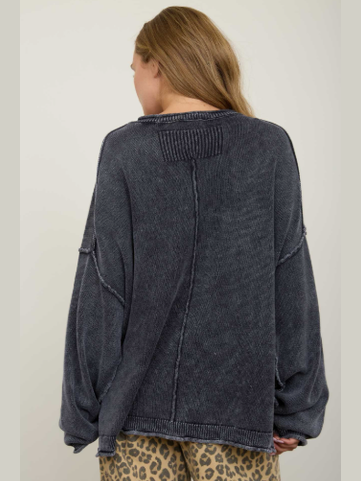 A woman viewed from the back wearing an oversized dark charcoal gray knit sweater with a textured, washed finish and leopard-print pants against a plain off-white background.
