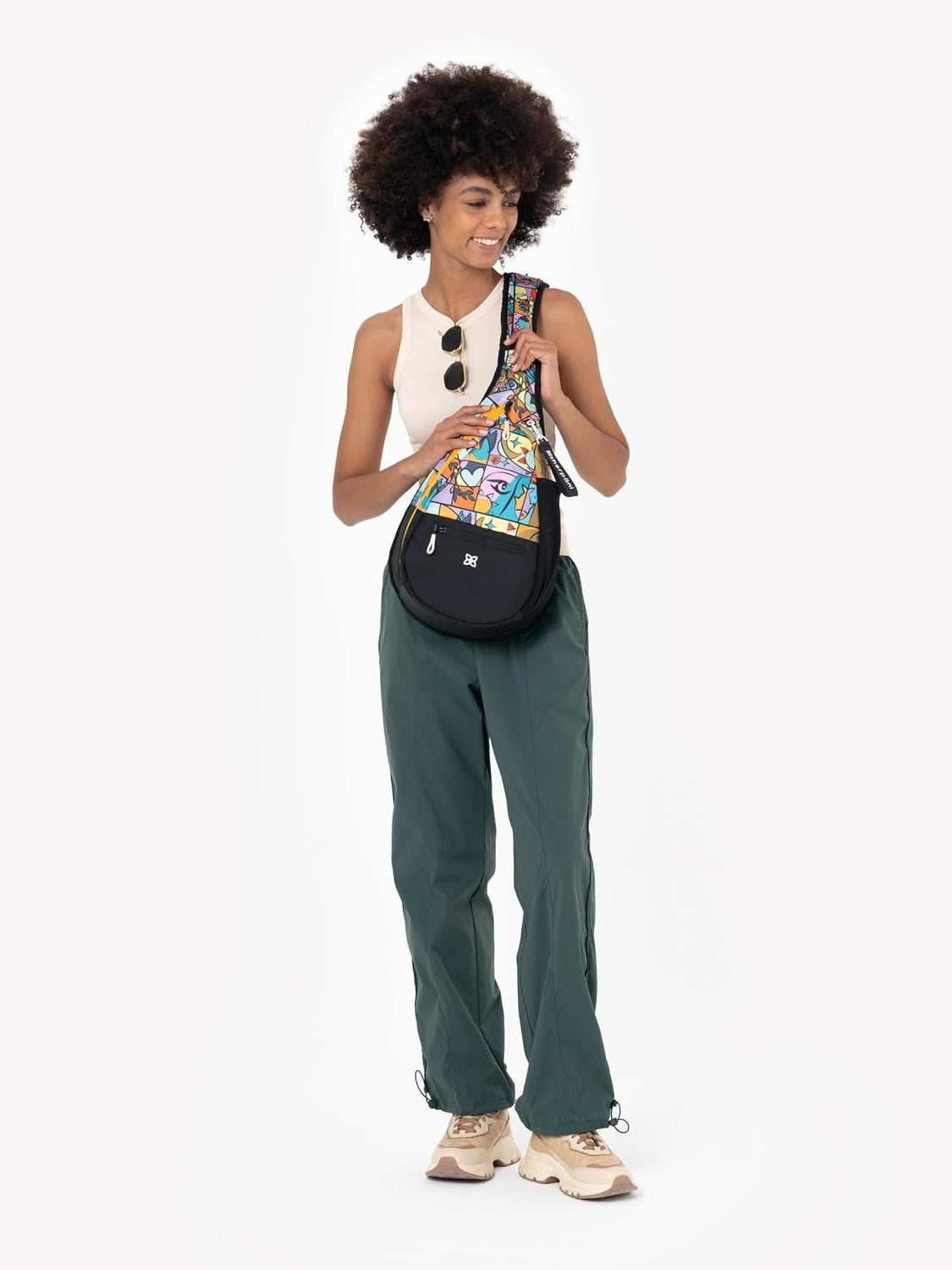 A woman with an afro hairstyle models a black crescent-shaped crossbody bag featuring a colorful, patterned front panel. She is wearing a white sleeveless top, dark green wide-leg pants, and beige sneakers against a plain white studio background.