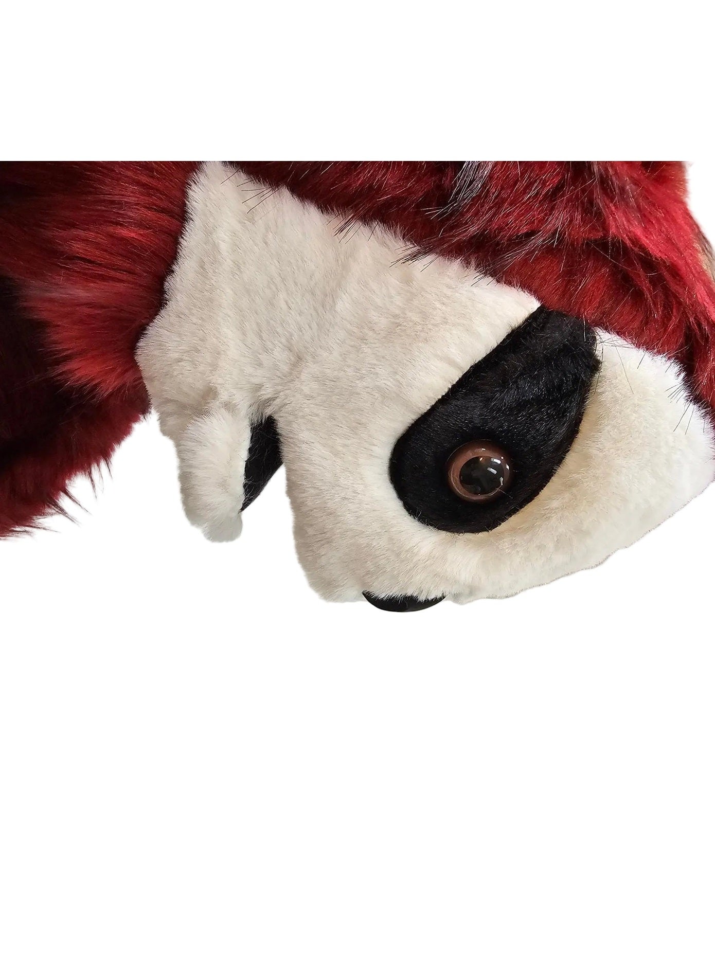 A close-up side view of a plush animal character with a white face, black eye patch, and deep red furry body, photographed indoors against a white paneled wall.