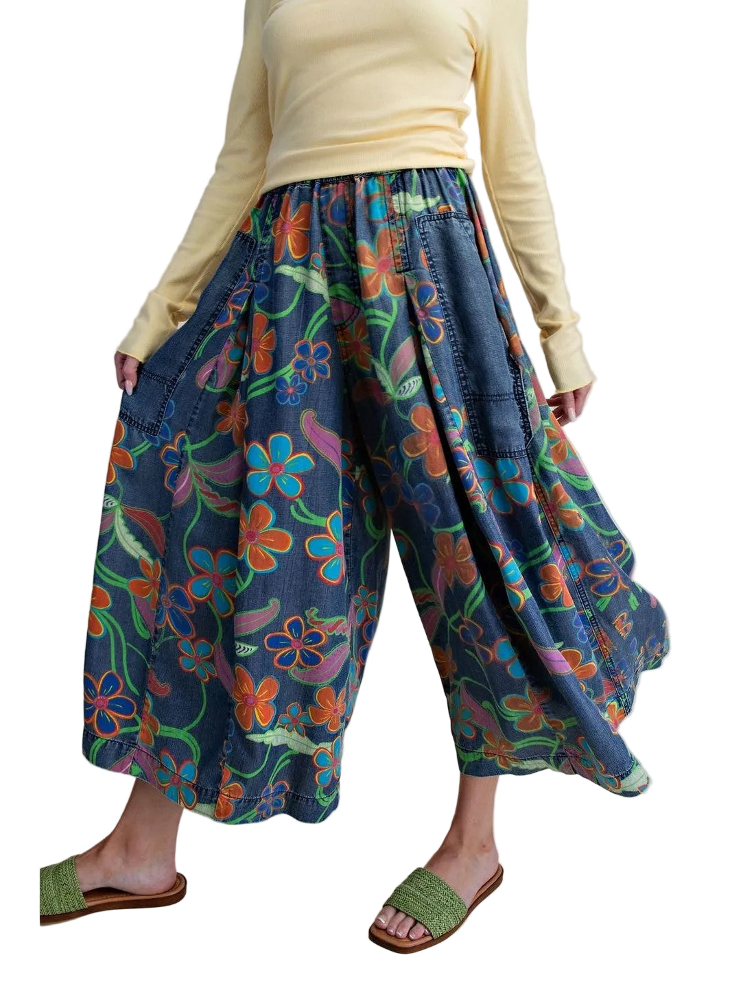 A person wearing loose, wide-leg cropped pants with a colorful floral print on a blue denim-like base, paired with a pale yellow long-sleeve top and green slide sandals, against a plain beige studio background.
