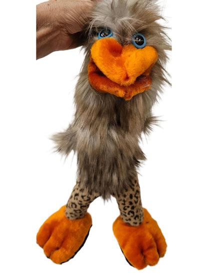 A plush ostrich puppet or toy with gray-brown fluffy feathers, a bright orange beak and feet, leopard-print legs, and blue eyes, shown with a hand holding its head against a white paneled wall.
