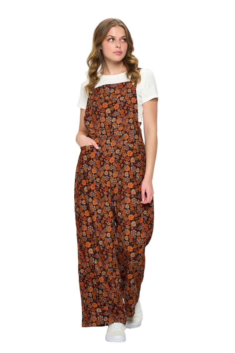A woman modeling a brown floral print overall jumpsuit with a white t-shirt underneath, standing against a plain white studio background.