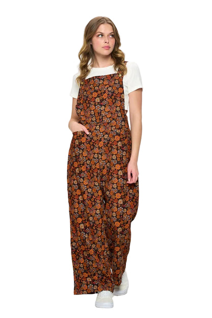 A woman modeling a brown floral print overall jumpsuit with a white t-shirt underneath, standing against a plain white studio background.
