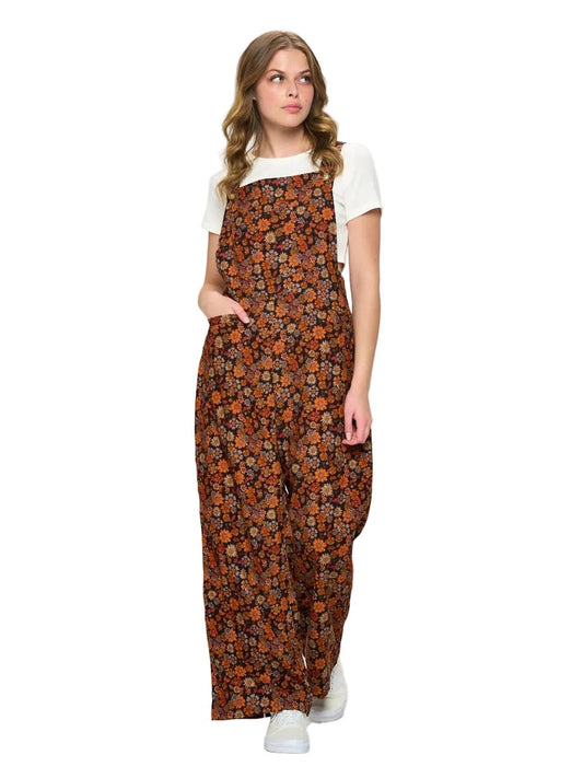 A woman modeling a brown floral print overall jumpsuit with a white t-shirt underneath, standing against a plain white studio background.