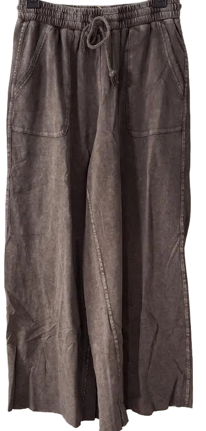 A pair of wide-leg cropped pants in a washed, faded brown or taupe color, hanging on a black hanger against a white paneled door. The pants feature an elastic waistband with a drawstring, side pockets, and a raw, cropped hem.