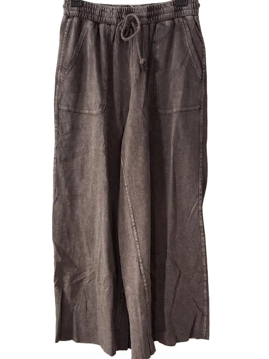A pair of wide-leg cropped pants in a washed, faded brown or taupe color, hanging on a black hanger against a white paneled door. The pants feature an elastic waistband with a drawstring, side pockets, and a raw, cropped hem.