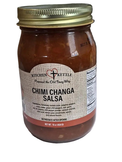 A glass jar of Kitchen Kettle Chimi Changa Salsa with a gold metal lid and a beige label, set on a white surface with a kitchen countertop in the background.