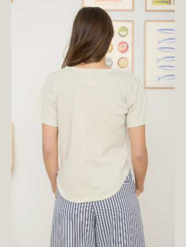 A woman viewed from behind wearing a light cream-colored short-sleeve top and blue-and-white striped pants, standing against a white wall with framed art.
