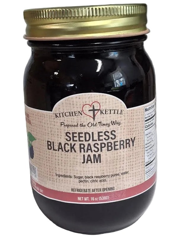 A jar of Kitchen Kettle Seedless Black Raspberry Jam with a gold metal lid and a beige label, set on a white surface with a gray background.