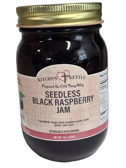 A jar of Kitchen Kettle Seedless Black Raspberry Jam with a gold metal lid and a beige label, set on a white surface with a gray background.