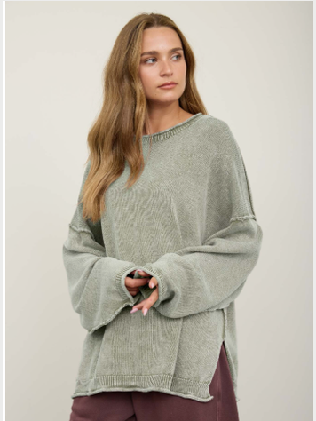 A woman wearing a light sage green oversized ribbed knit sweater with wide bell sleeves, paired with dark plum pants, against a plain light beige studio background.