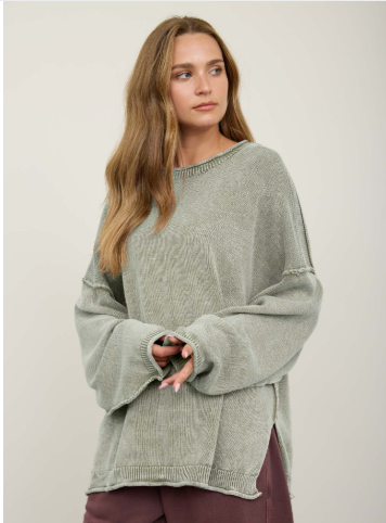 A woman wearing a light sage green oversized ribbed knit sweater with wide bell sleeves, paired with dark plum pants, against a plain light beige studio background.