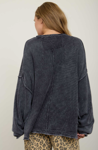 A woman viewed from the back wearing an oversized dark charcoal gray knit sweater with a textured, washed finish and leopard-print pants against a plain off-white background.