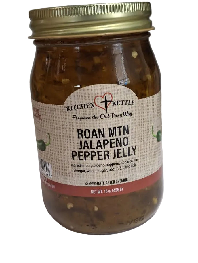A glass jar of Kitchen Kettle Roan Mtn Jalapeño Pepper Jelly with a gold metal lid and a beige burlap-style label, photographed on a white surface with a gray background.