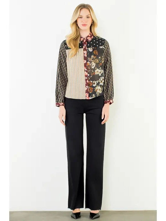 A woman stands against a plain white wall wearing a long-sleeve collared blouse with mixed patterns: vertical beige and black stripes on the left, floral print on the right, and a black-and-white gingham pattern on the sleeves, paired with black wide-leg pants and black shoes.