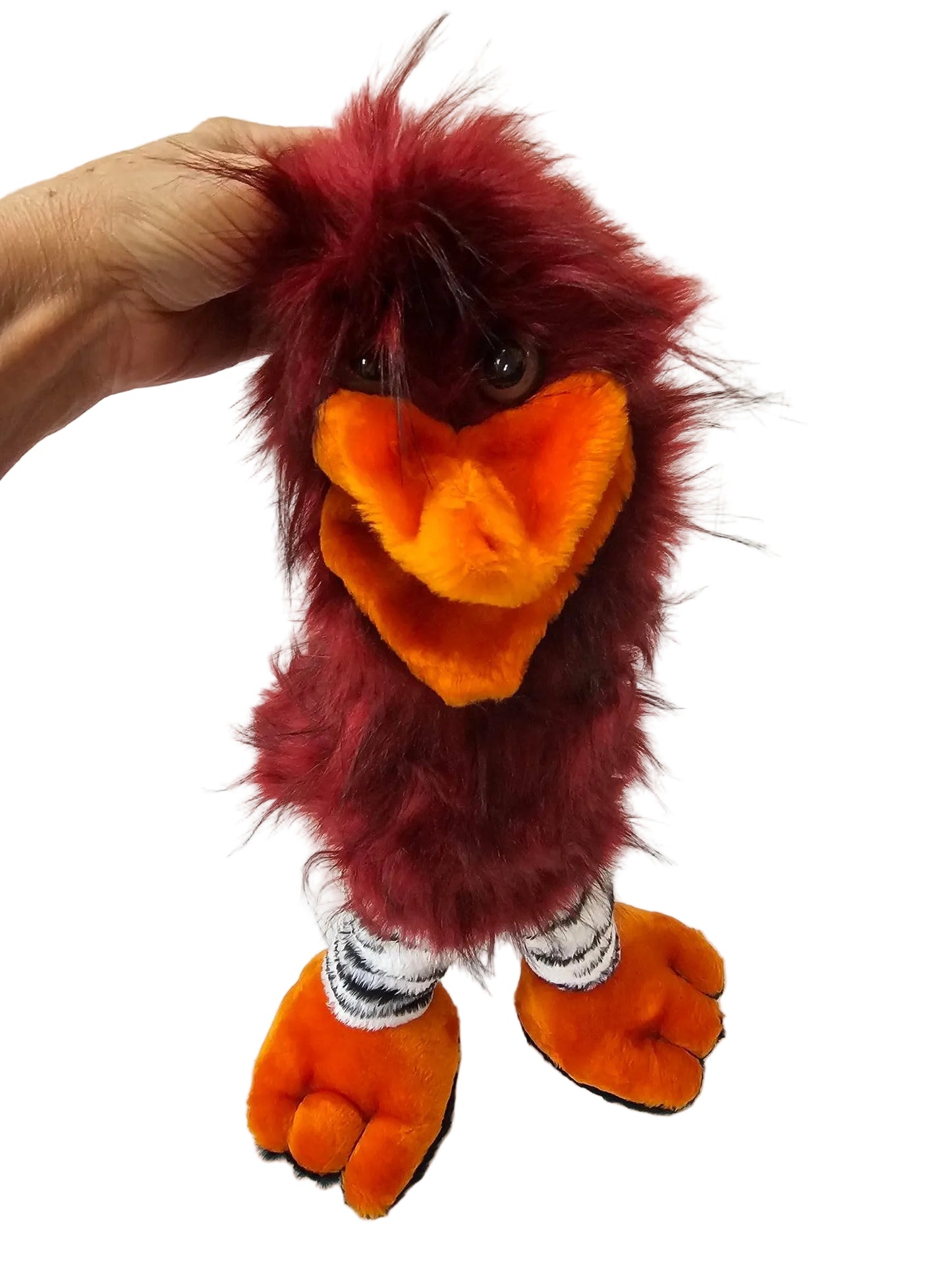 A plush bird puppet or hand puppet with dark red fur, a bright orange beak, and orange feet, being held by a hand against a white paneled wall.