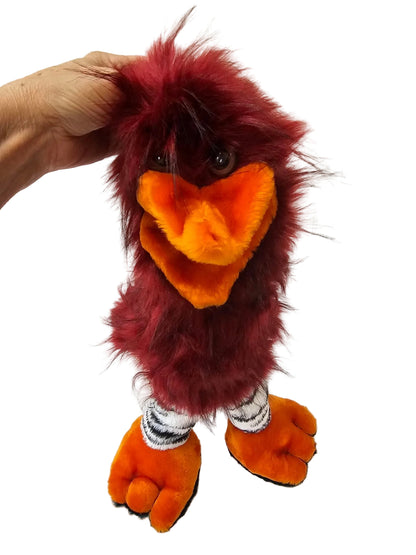 A plush bird puppet or hand puppet with dark red fur, a bright orange beak, and orange feet, being held by a hand against a white paneled wall.