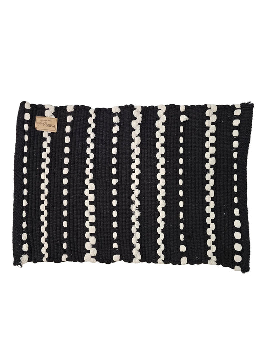 A rectangular black woven rug with vertical white dotted stripes, laid flat on a light gray surface. A small tan paper label is attached near the top left corner.