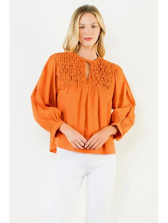 A woman modeling a burnt orange blouse with a textured crochet-style yoke and keyhole neckline, paired with white pants, against a plain white studio background.