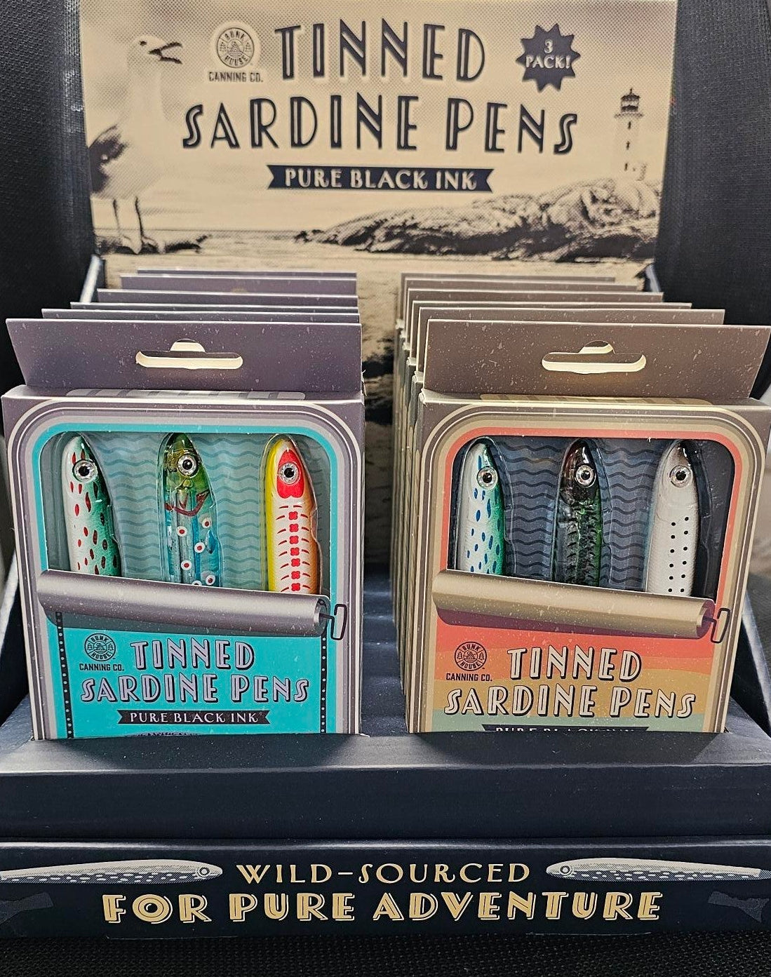 A boxed set of tinned sardines and tinned paddle fish packaged in decorative tin-style boxes, displayed on a dark textured surface with a black tray holding a card that reads 'Tinned Sardine Pens' and 'Wild Pouched.'.