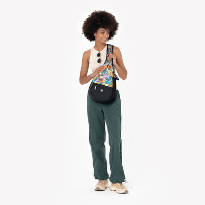 A woman with an afro hairstyle models a black crescent-shaped crossbody bag featuring a colorful, patterned front panel. She is wearing a white sleeveless top, dark green wide-leg pants, and beige sneakers against a plain white studio background.
