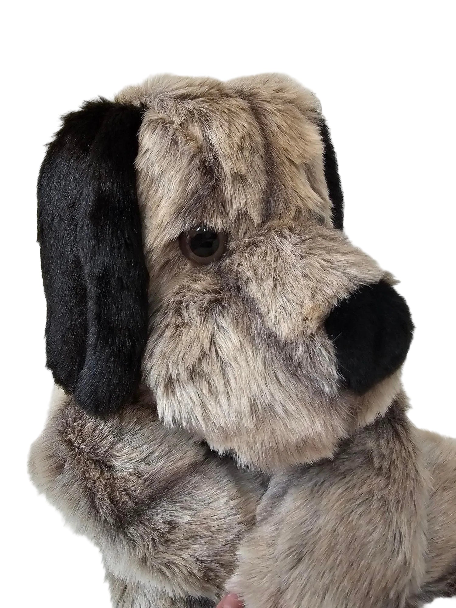 A close-up of a plush dog toy or stuffed animal head made from faux fur, with a black nose and ears, a brown eye, and a gray and beige fur body, photographed against a white paneled background.
