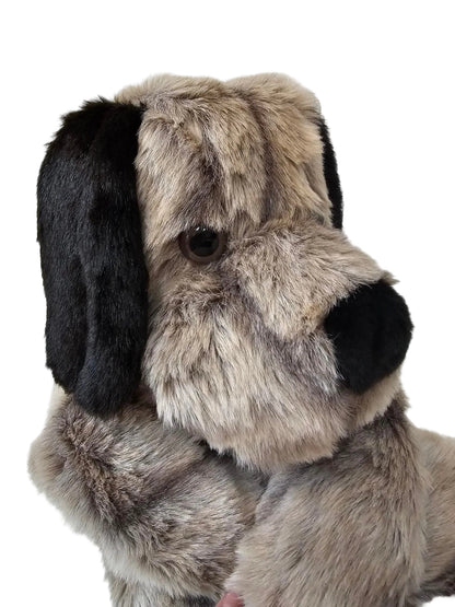 A close-up of a plush dog toy or stuffed animal head made from faux fur, with a black nose and ears, a brown eye, and a gray and beige fur body, photographed against a white paneled background.