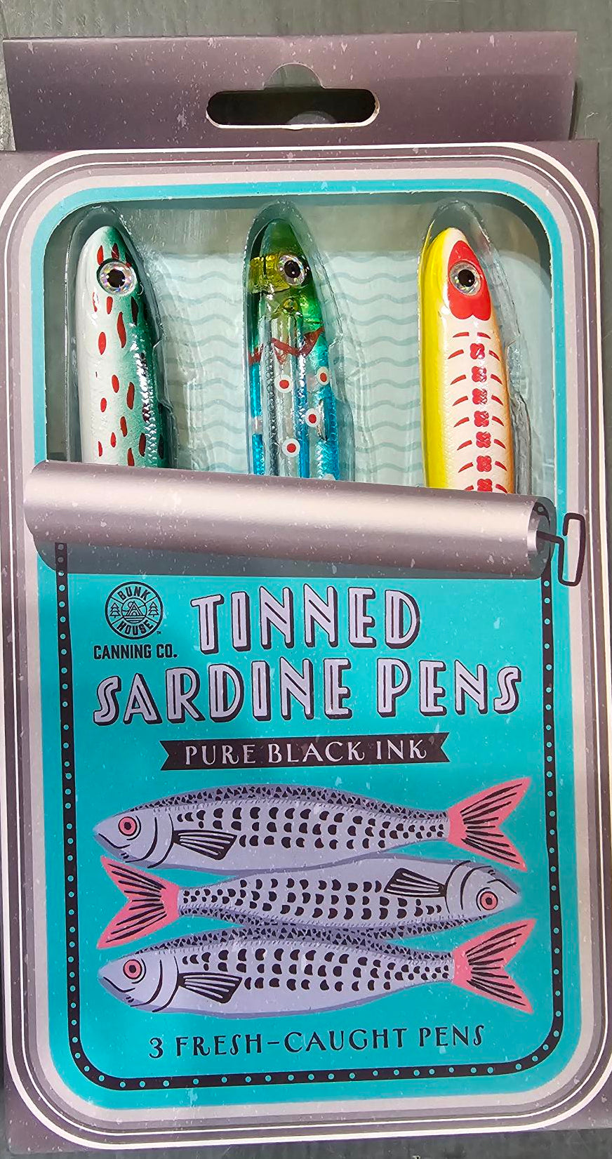 A tin of three tinned sardine pens on a gray surface. The packaging features a turquoise label with illustrations of sardines and the text 'TINNED SARDINE PENS' and 'PURE BLACK INK'.