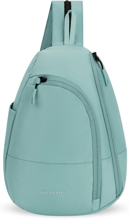 A light mint green sling backpack by Sherpani, featuring a single shoulder strap, a front zippered pocket, and a curved lower body, shown against a plain white background.