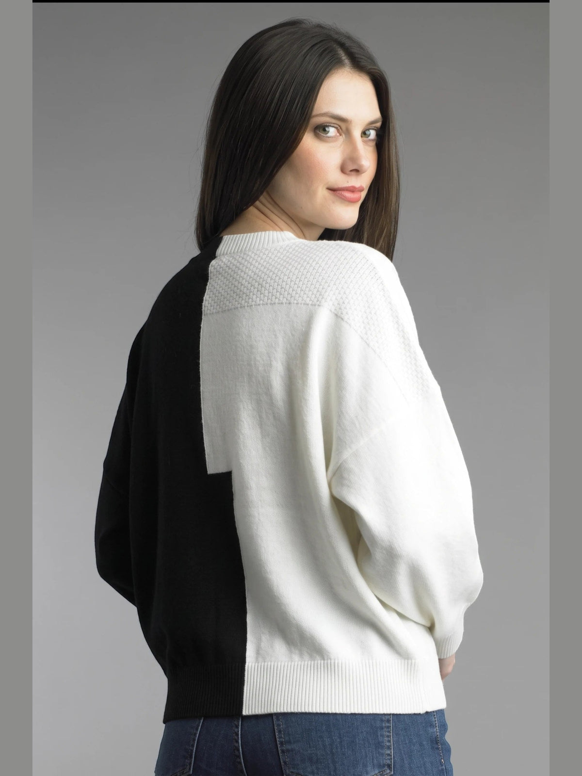 A woman wearing a color-block knit sweater with a white front panel and black side/back panel, paired with blue jeans, photographed against a plain gray studio background.