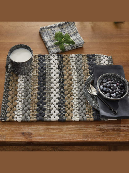 A woven placemat with vertical stripes in gray, beige, white, and black sits on a wooden table, styled with a speckled mug of milk, a bowl of blueberries, a spoon, and a folded napkin.