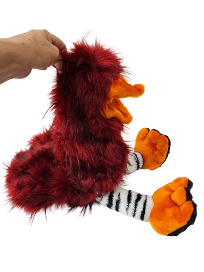 A plush toy bird with shaggy dark red fur, a bright orange beak, orange feet with black trim, and striped black-and-white legs, shown on a white surface with a hand holding the top of its head.