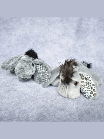 A plush stuffed animal toy shaped like a gray elephant lying on a soft light gray fabric background. The toy has dark brown faux fur on its head and ears, a black-and-white checkered necktie, and a light blue patterned inner ear.