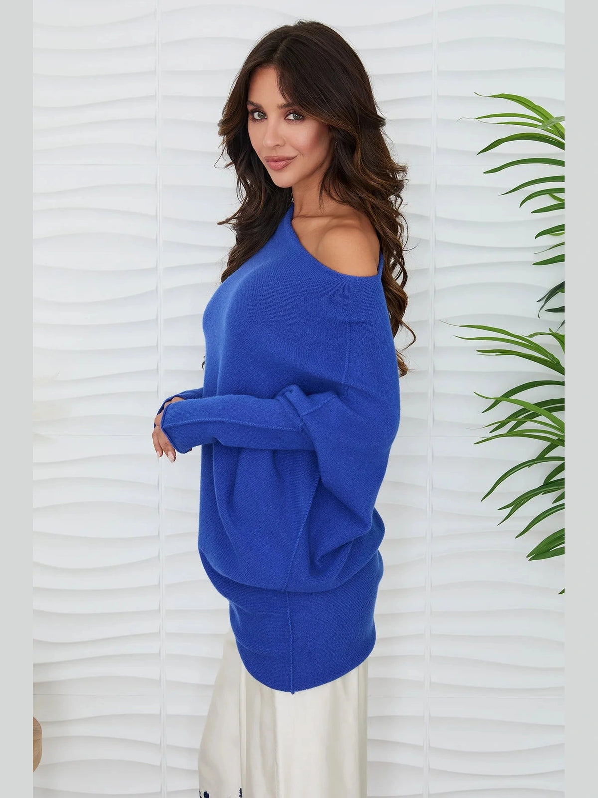 A woman wearing a bright royal blue off-the-shoulder knit sweater, paired with a cream-colored skirt, standing against a white wavy wall with a green plant on the right.