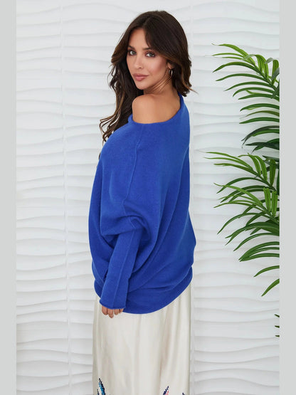 A woman wearing a vibrant royal blue off-the-shoulder knit sweater and a cream-colored skirt, standing against a white wavy wall with a green plant on the right.