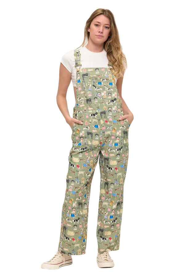 A woman modeling a pair of sage green bib overalls with a dense multicolor cartoon character print, styled over a white t-shirt and cream sneakers against a plain white studio background.