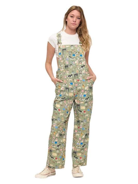 A woman modeling a pair of sage green bib overalls with a dense multicolor cartoon character print, styled over a white t-shirt and cream sneakers against a plain white studio background.