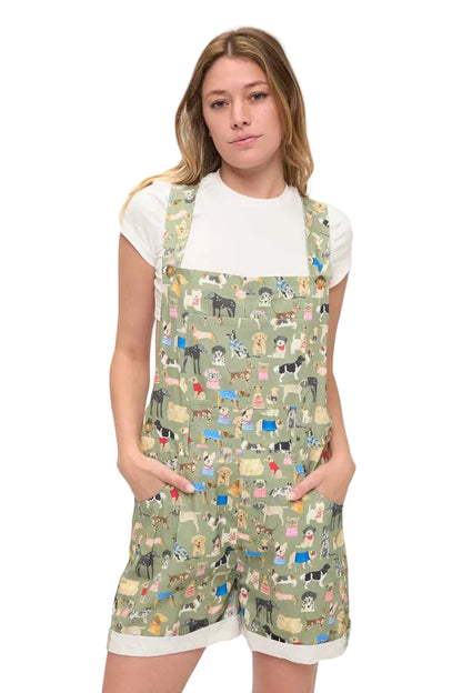 A young woman modeling a white t-shirt and sage green overall shorts covered in a colorful cartoon dog pattern, set against a plain white studio background.