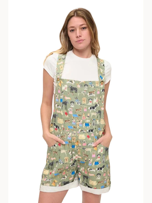 A young woman modeling a white t-shirt and sage green overall shorts covered in a colorful cartoon dog pattern, set against a plain white studio background.
