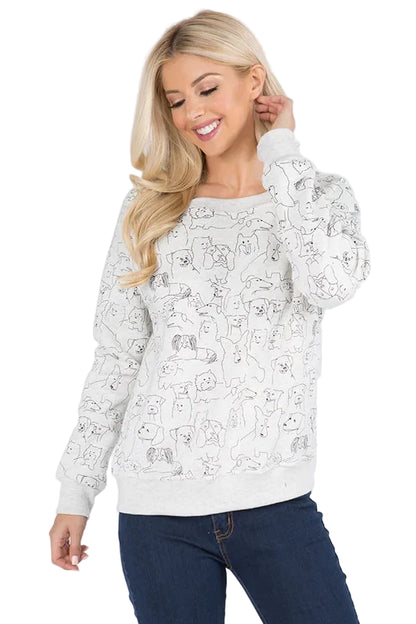 A blonde woman wearing a white long-sleeve crewneck sweatshirt with a black line art pattern of cartoon dogs, paired with dark blue jeans, against a light gray studio background.