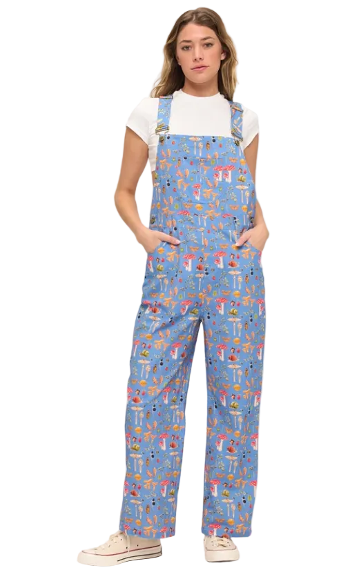 A woman modeling light blue overall-style coveralls with a colorful all-over print, worn over a white short-sleeve T-shirt and paired with beige sneakers, against a plain light background.