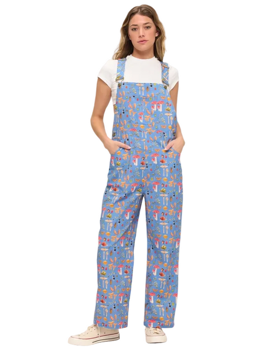 A woman modeling light blue overall-style coveralls with a colorful all-over print, worn over a white short-sleeve T-shirt and paired with beige sneakers, against a plain light background.