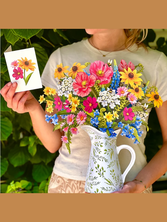 A person outdoors holds a white floral pitcher filled with a colorful bouquet of flowers, including pink, yellow, blue, and white blossoms. The person also holds up a small card showing a matching flower illustration.