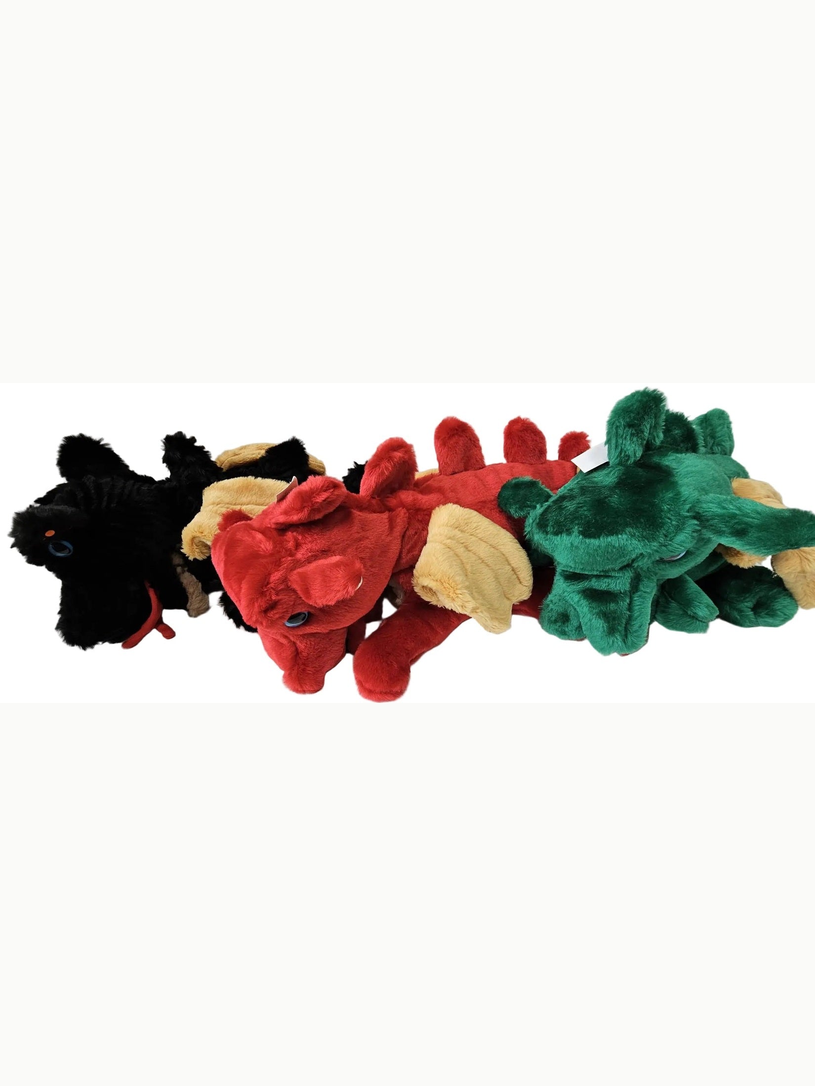 Three plush dragon toys lined up on a white surface against a white paneled wall. The dragons are black, red, and green, each with tan wings and small red accents.
