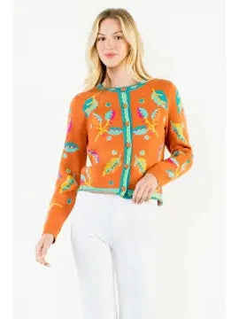 A blonde model wearing a bright orange knit cardigan with colorful embroidered floral motifs in green, yellow, and pink, paired with white pants, against a plain white studio background.