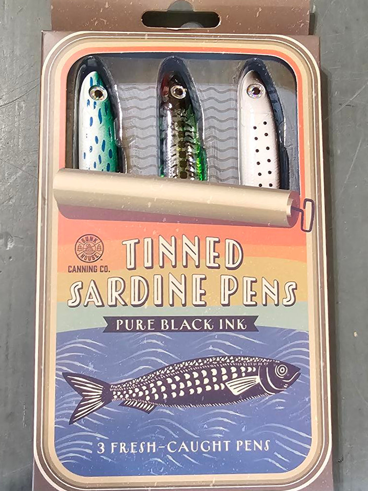 A boxed set of three tinned sardine pens fishing lures on a gray metal surface. The packaging features a blue and orange design with an illustration of a sardine and the text 'Tinned Sardine Pens' and 'Three Black Ink'.
