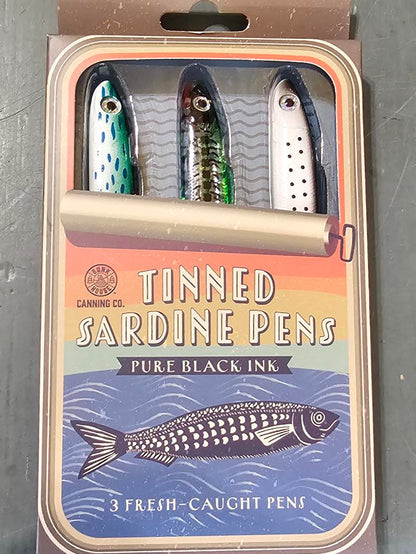 A boxed set of three tinned sardine pens fishing lures on a gray metal surface. The packaging features a blue and orange design with an illustration of a sardine and the text 'Tinned Sardine Pens' and 'Three Black Ink'.