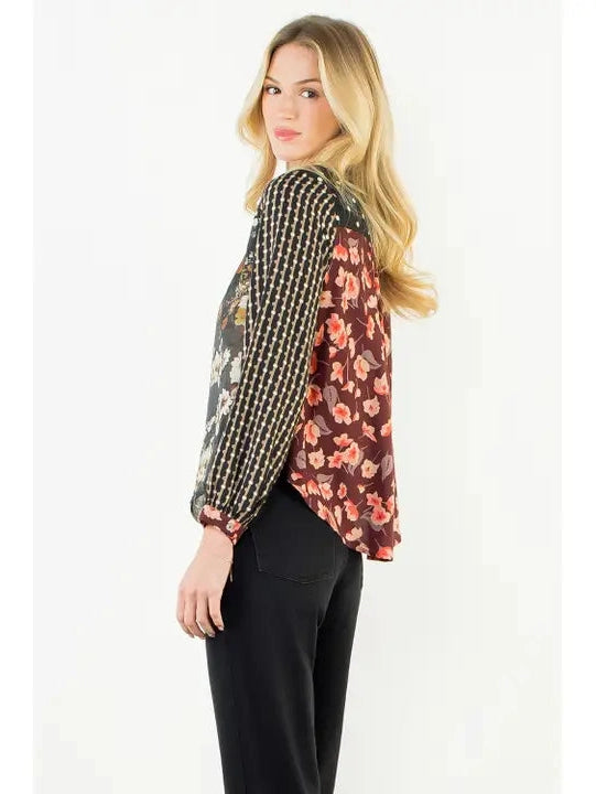 A woman modeling a long-sleeve blouse with a mixed print design, featuring a black and white checkered pattern on one side and a dark red floral pattern on the other, paired with black pants against a plain white background.