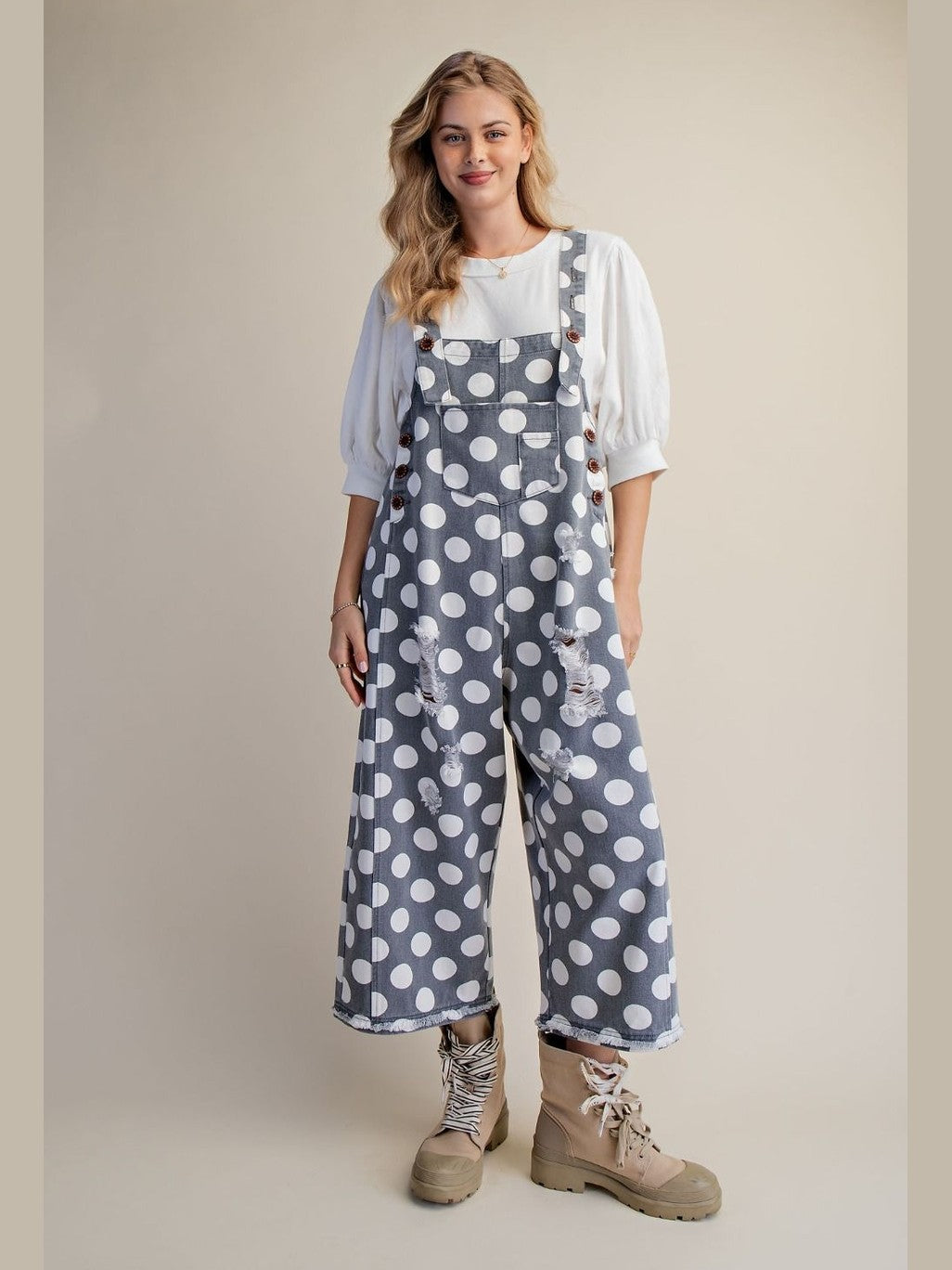 A woman modeling slate blue wide-leg overall with white polka dots, styled with a white puff-sleeve top and beige lace-up boots, against a solid beige studio background.