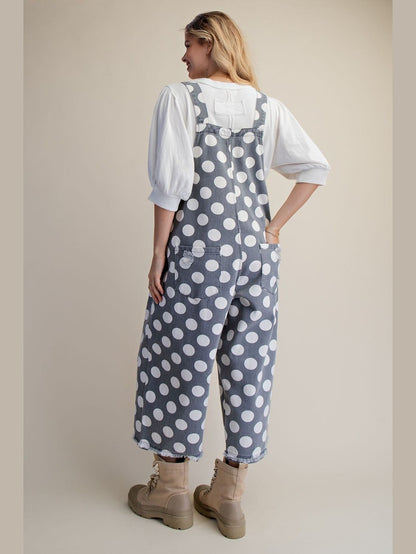 A woman shown from the back wearing slate blue denim-style overalls with a white polka dot pattern, layered over a white puff-sleeve top and paired with beige combat boots, against a solid beige studio background.